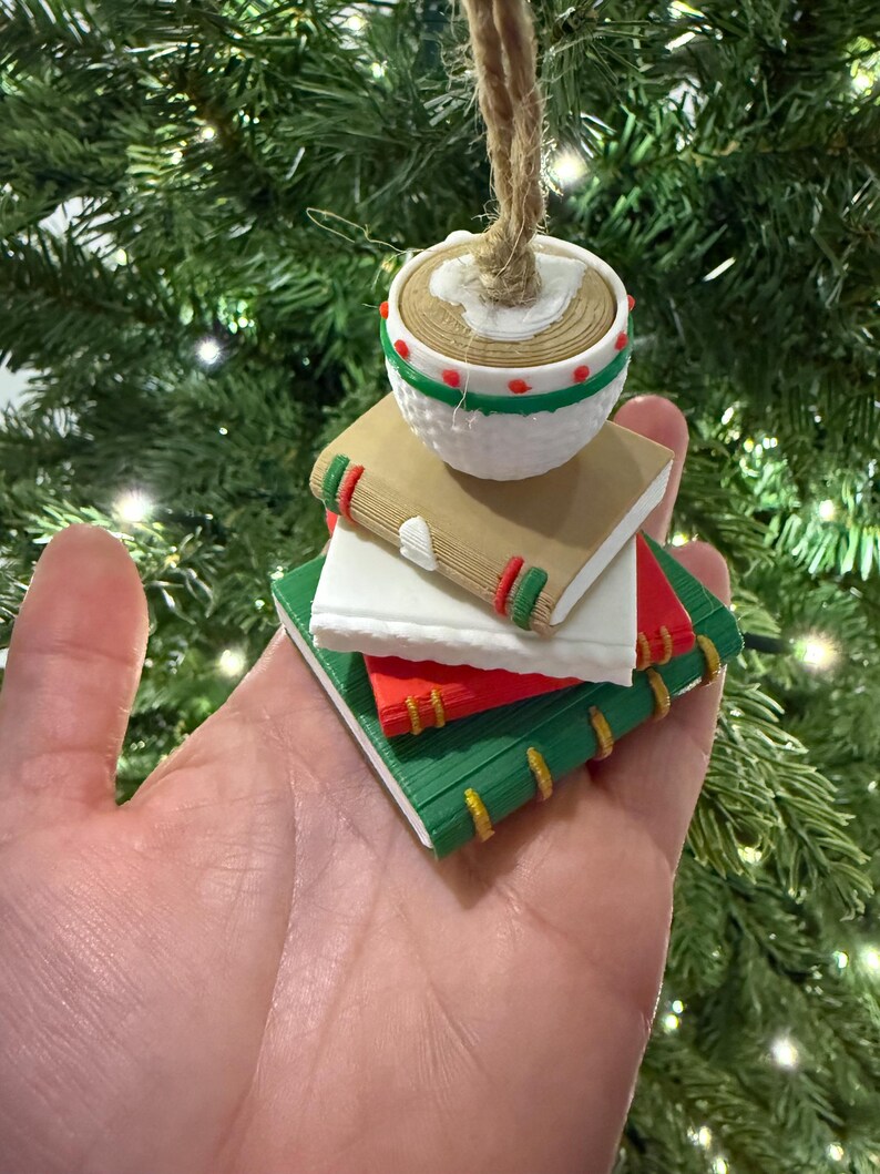 K&ouml;nnte beinhalten: Ein Weihnachtsbaumschmuck in Form eines Stapels Miniaturb&uuml;cher, gekr&ouml;nt von einem runden, strukturierten Ornament. Die B&uuml;cher sind in Braun-, Wei&szlig;-, Rot- und Gr&uuml;nt&ouml;nen gehalten und weisen dekorative Details auf. Eine Schnurschlaufe ist zum Aufh&auml;ngen befestigt.