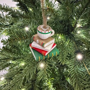 K&ouml;nnte beinhalten: Ein Weihnachtsbaumschmuck mit einer Kaffeetasse auf einem Stapel aus drei B&uuml;chern. Die B&uuml;cher sind rot, wei&szlig; und gr&uuml;n mit goldenen Buchr&uuml;cken. Die Tasse ist wei&szlig; mit gr&uuml;nen und roten Details und enth&auml;lt eine braune Fl&uuml;ssigkeit.