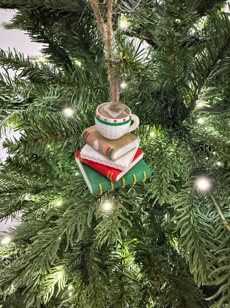 K&ouml;nnte beinhalten: Ein Weihnachtsbaumschmuck mit einer Kaffeetasse auf einem Stapel aus drei B&uuml;chern. Die B&uuml;cher sind rot, wei&szlig; und gr&uuml;n mit goldenen Buchr&uuml;cken. Die Tasse ist wei&szlig; mit gr&uuml;nen und roten Details und enth&auml;lt eine braune Fl&uuml;ssigkeit.