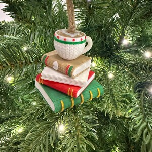 K&ouml;nnte beinhalten: Ein Weihnachtsbaumschmuck mit einer Kaffeetasse auf einem Stapel aus drei B&uuml;chern. Die B&uuml;cher sind rot, wei&szlig; und gr&uuml;n mit goldenen Buchr&uuml;cken. Die Tasse ist wei&szlig; mit roten und gr&uuml;nen Akzenten. Der Schmuck h&auml;ngt an einer nat&uuml;rlichen Schnur.