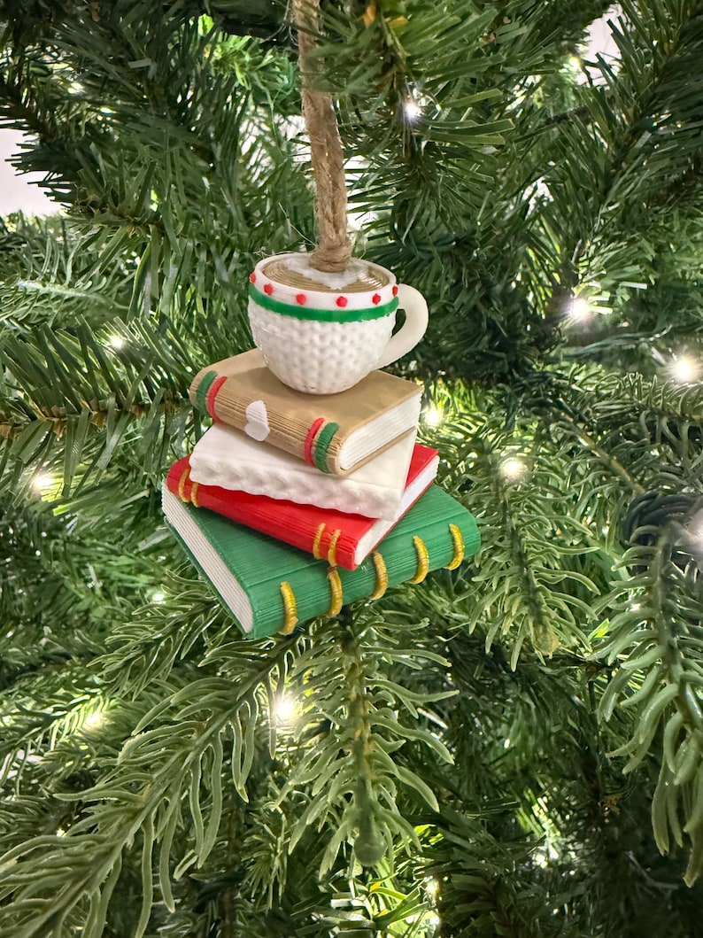 K&ouml;nnte beinhalten: Ein Weihnachtsbaumschmuck mit einer Kaffeetasse auf einem Stapel aus drei B&uuml;chern. Die B&uuml;cher sind rot, wei&szlig; und gr&uuml;n mit goldenen Buchr&uuml;cken. Die Tasse ist wei&szlig; mit roten und gr&uuml;nen Akzenten. Der Schmuck h&auml;ngt an einer nat&uuml;rlichen Schnur.