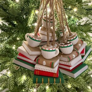 K&ouml;nnte beinhalten: Weihnachtsbaumschmuck mit wei&szlig;en Tassen mit rot-gr&uuml;nen Verzierungen, die auf B&uuml;chern liegen. Die B&uuml;cher sind in verschiedenen Farben gehalten, darunter Rot, Gr&uuml;n und Braun, und werden mit Schnur aufgeh&auml;ngt. Festlich und dekorativ.