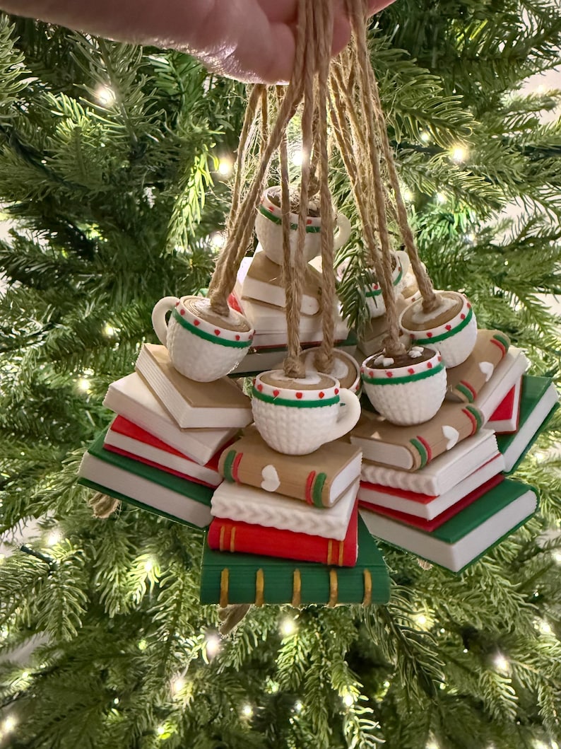 K&ouml;nnte beinhalten: Weihnachtsbaumschmuck mit wei&szlig;en Tassen mit rot-gr&uuml;nen Verzierungen, die auf B&uuml;chern liegen. Die B&uuml;cher sind in verschiedenen Farben gehalten, darunter Rot, Gr&uuml;n und Braun, und werden mit Schnur aufgeh&auml;ngt. Festlich und dekorativ.