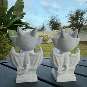 May include: Two gray gargoyle statues with textured surfaces, each featuring bat-like wings, pointed ears, and a curled tail. The statues are positioned on a dark surface against a backdrop of greenery and a cloudy sky.