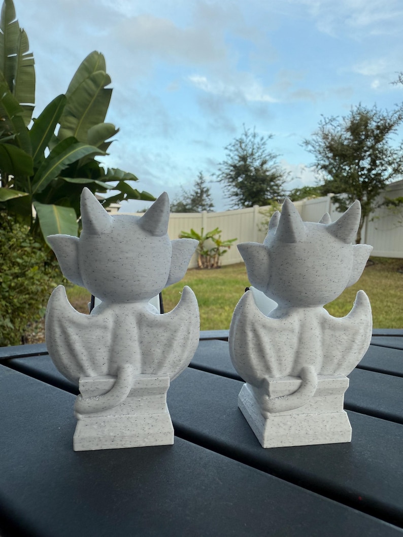 May include: Two gray gargoyle statues with textured surfaces, each featuring bat-like wings, pointed ears, and a curled tail. The statues are positioned on a dark surface against a backdrop of greenery and a cloudy sky.