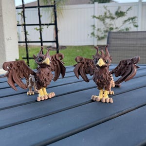 May include: Two brown griffin figurines with outstretched wings, yellow feet, and detailed feather textures. The mythical creatures stand on a dark gray surface, showcasing their intricate design and craftsmanship.