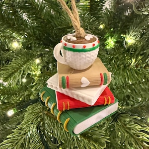K&ouml;nnte beinhalten: Ein Weihnachtsbaumschmuck mit einer wei&szlig;en Tasse hei&szlig;er Schokolade mit roten und gr&uuml;nen Akzenten, die auf drei bunten B&uuml;chern gestapelt ist. Die B&uuml;cher sind gr&uuml;n, rot und wei&szlig;, mit goldfarbenen Buchr&uuml;cken. Der Schmuck h&auml;ngt an einer Schnur.