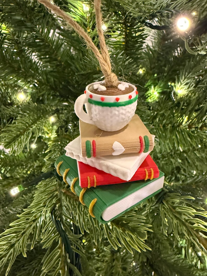 K&ouml;nnte beinhalten: Ein Weihnachtsbaumschmuck mit einer wei&szlig;en Tasse hei&szlig;er Schokolade mit roten und gr&uuml;nen Akzenten, die auf drei bunten B&uuml;chern gestapelt ist. Die B&uuml;cher sind gr&uuml;n, rot und wei&szlig;, mit goldfarbenen Buchr&uuml;cken. Der Schmuck h&auml;ngt an einer Schnur.