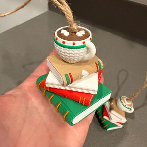 K&ouml;nnte beinhalten: Ein dekorativer Ornament mit einem Stapel Miniaturb&uuml;cher in Gr&uuml;n, Rot und Beige, gekr&ouml;nt von einer wei&szlig;en Tasse Kakao. Die Tasse hat einen Henkel und ist mit roten und gr&uuml;nen Akzenten verziert. Eine Schnurschlaufe ist befestigt.