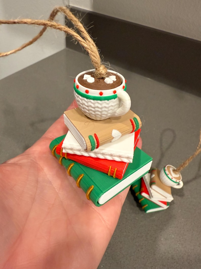 K&ouml;nnte beinhalten: Ein dekorativer Ornament mit einem Stapel Miniaturb&uuml;cher in Gr&uuml;n, Rot und Beige, gekr&ouml;nt von einer wei&szlig;en Tasse Kakao. Die Tasse hat einen Henkel und ist mit roten und gr&uuml;nen Akzenten verziert. Eine Schnurschlaufe ist befestigt.