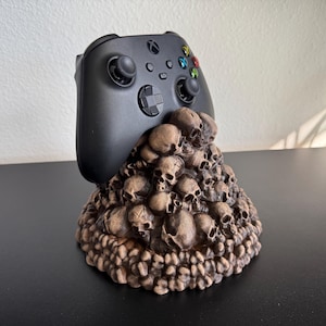 May include: A black Xbox controller rests on a brown skull-shaped stand. The stand is intricately detailed with numerous small skulls. The controller has colored buttons and directional pad. The stand is on a black surface.