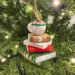 K&ouml;nnte beinhalten: Weihnachtsbaumschmuck mit einem Stapel B&uuml;cher, gekr&ouml;nt von einer Tasse hei&szlig;er Schokolade. Die B&uuml;cher sind rot, gr&uuml;n und beige mit wei&szlig;en Seiten. Die Tasse ist wei&szlig; mit gr&uuml;nen und roten Verzierungen und einer braunen Fl&uuml;ssigkeit.