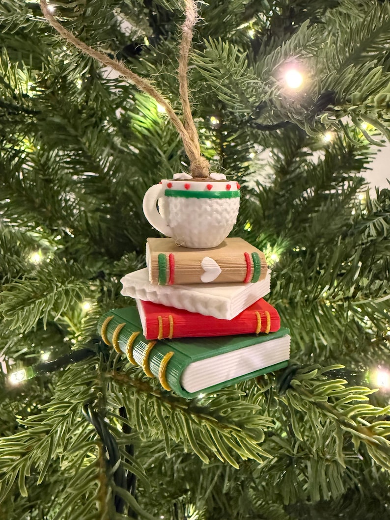 K&ouml;nnte beinhalten: Weihnachtsbaumschmuck mit einem Stapel B&uuml;cher, gekr&ouml;nt von einer Tasse hei&szlig;er Schokolade. Die B&uuml;cher sind rot, gr&uuml;n und beige mit wei&szlig;en Seiten. Die Tasse ist wei&szlig; mit gr&uuml;nen und roten Verzierungen und einer braunen Fl&uuml;ssigkeit.