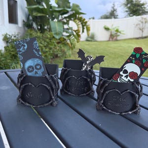 May include: Three black gothic-style holders. Each holder features a different inscription: "Smut", "Trigger Warning", and "Dark Romance". Each holder contains a decorative card with a skull or bat design.