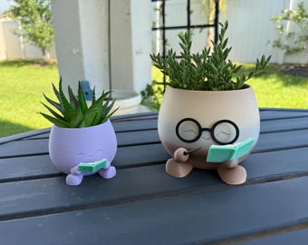 May include: Two whimsical planters, one lavender and one beige, each holding greenery. The planters are designed with faces, glasses, and tiny books, creating a charming, playful aesthetic. The planters are sitting on a dark gray table.