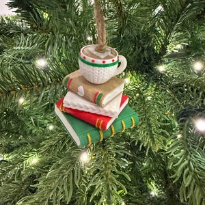 K&ouml;nnte beinhalten: Ein Weihnachtsbaumschmuck mit einer Kaffeetasse auf einem Stapel aus drei B&uuml;chern. Die B&uuml;cher sind rot, wei&szlig; und gr&uuml;n mit goldenen Buchr&uuml;cken. Die wei&szlig;e Tasse hat rote und gr&uuml;ne Akzente und einen braunen Faden zum Aufh&auml;ngen.