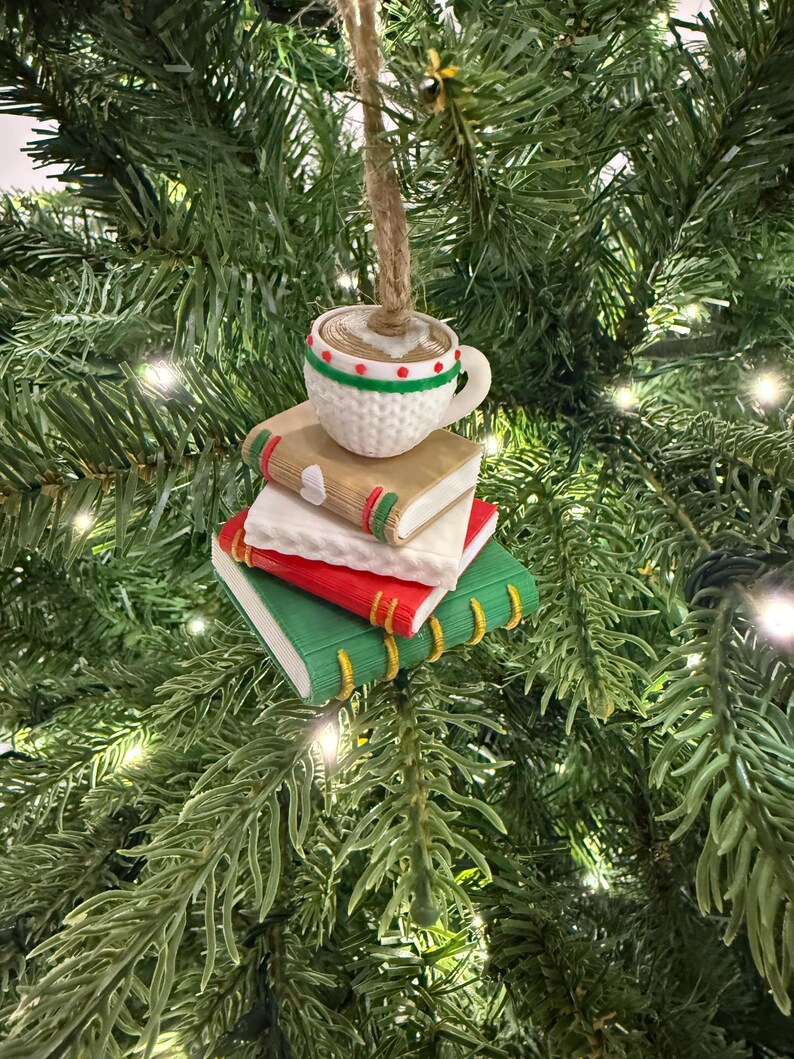 K&ouml;nnte beinhalten: Ein Weihnachtsbaumschmuck mit einer Kaffeetasse auf einem Stapel aus drei B&uuml;chern. Die B&uuml;cher sind rot, wei&szlig; und gr&uuml;n mit goldenen Buchr&uuml;cken. Die wei&szlig;e Tasse hat rote und gr&uuml;ne Akzente und einen braunen Faden zum Aufh&auml;ngen.