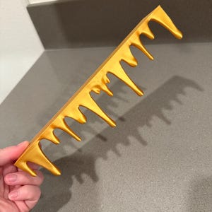 Peut inclure: Un peigne dor&eacute; au design unique. Les dents du peigne ont un effet d&eacute;goulinant, cr&eacute;ant une apparence artistique et accrocheuse. Le peigne est tenu dans une main sur un comptoir gris.