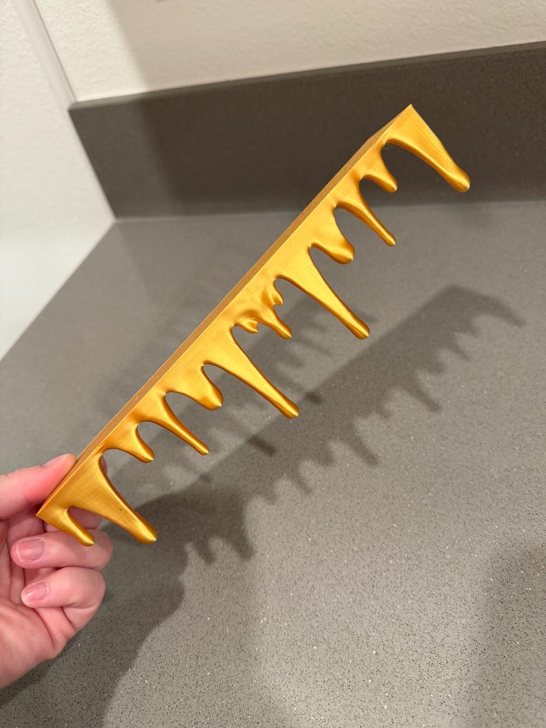 Peut inclure: Un peigne dor&eacute; au design unique. Les dents du peigne ont un effet d&eacute;goulinant, cr&eacute;ant une apparence artistique et accrocheuse. Le peigne est tenu dans une main sur un comptoir gris.