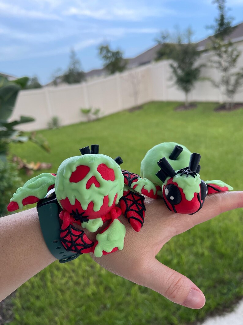 Puede incluir: Accesorio decorativo verde y rojo para smartwatch. Presenta un dise&ntilde;o de calavera con un efecto verde goteante, detalles rojos y una telara&ntilde;a. La correa del reloj es verde oscuro.
