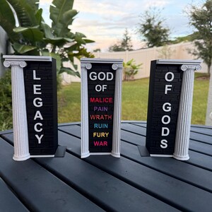 May include: Three black and white decorative pillars with text. The left pillar reads "LEGACY", the center pillar reads "GOD OF" with the words "MALICE, PAIN, WRATH, RUIN, FURY, WAR" below, and the right pillar reads "OF GODS".