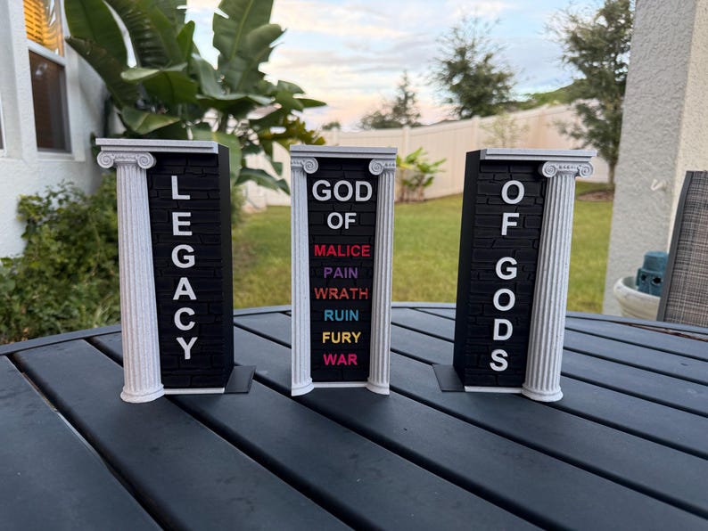 May include: Three black and white decorative pillars with text. The left pillar reads "LEGACY", the center pillar reads "GOD OF" with the words "MALICE, PAIN, WRATH, RUIN, FURY, WAR" below, and the right pillar reads "OF GODS".