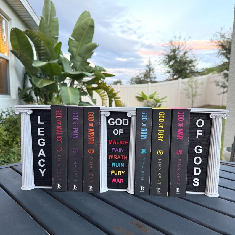 May include: A decorative white and black book display stand holding a collection of books. The books have black spines with titles like "God of Malice" and "God of War." The stand has the word "Legacy" and "Of Gods" on it.