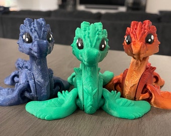 May include: Three small, articulated dragon figurines. The dragons are posed in a sitting position with their tails curled. The dragons are blue, green, and orange.