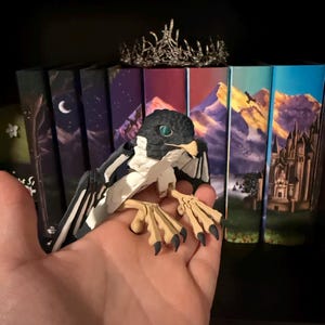 May include: A black and white figurine of a falcon with green eyes. The figurine is posed with its wings spread and talons extended. The figurine is sitting on a person's hand.