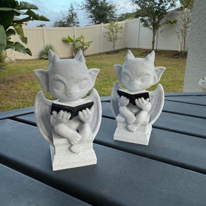 May include: Two gray gargoyle statues, each with wings and horns, are reading black books. The statues are sitting on a gray surface, with a white fence and green grass in the background.