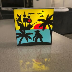 May include: A black pirate-themed decorative box featuring a silhouette of a pirate ship, palm trees, and a pirate on a tropical island. The background has a yellow sky, a red sun, and blue water. The box is made of black plastic.