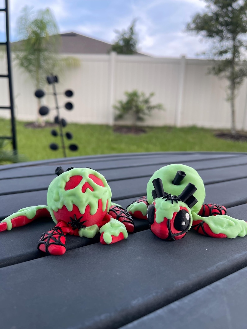 Puede incluir: Dos figuras de ara&ntilde;as con tem&aacute;tica de Halloween. Las ara&ntilde;as tienen cuerpos rojos con caras y patas verdes goteantes en forma de calavera. Tambi&eacute;n se ven telara&ntilde;as negras y peque&ntilde;as ara&ntilde;as negras. Las figuras est&aacute;n sobre una superficie gris oscuro.