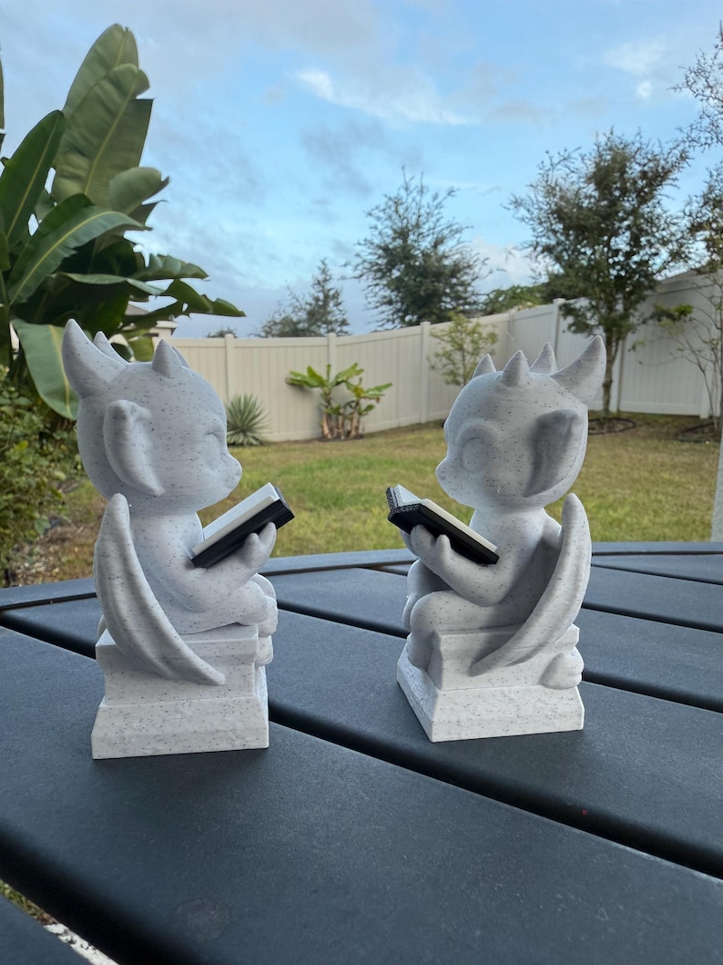 May include: Two gray dragon statues, each reading a black book. The statues have wings and sit on a rectangular base. The background shows a cloudy sky and green grass.