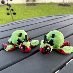 Puede incluir: Dos criaturas de arcilla hechas a mano, caprichosas, con cuerpos verdes, detalles rojos y negros, y antenas negras. Descansan sobre una superficie de madera gris, con un fondo verde borroso.