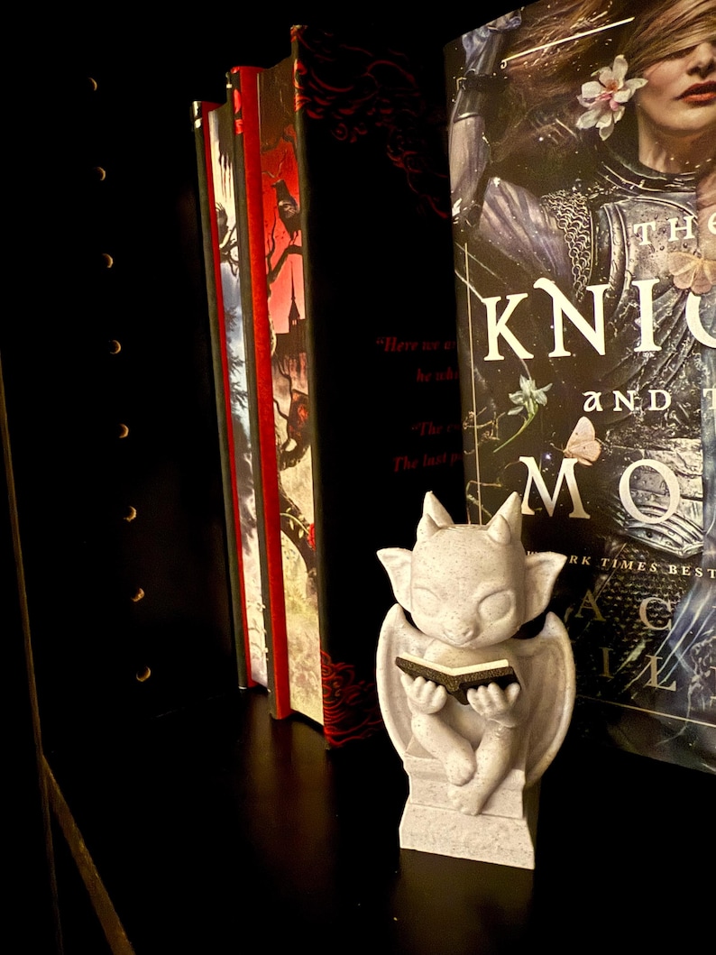 May include: A small, light gray gargoyle statue reading a book, positioned on a black shelf next to several books. The books have red and black covers. One book has the title "The Knights and the Moon" on the cover.