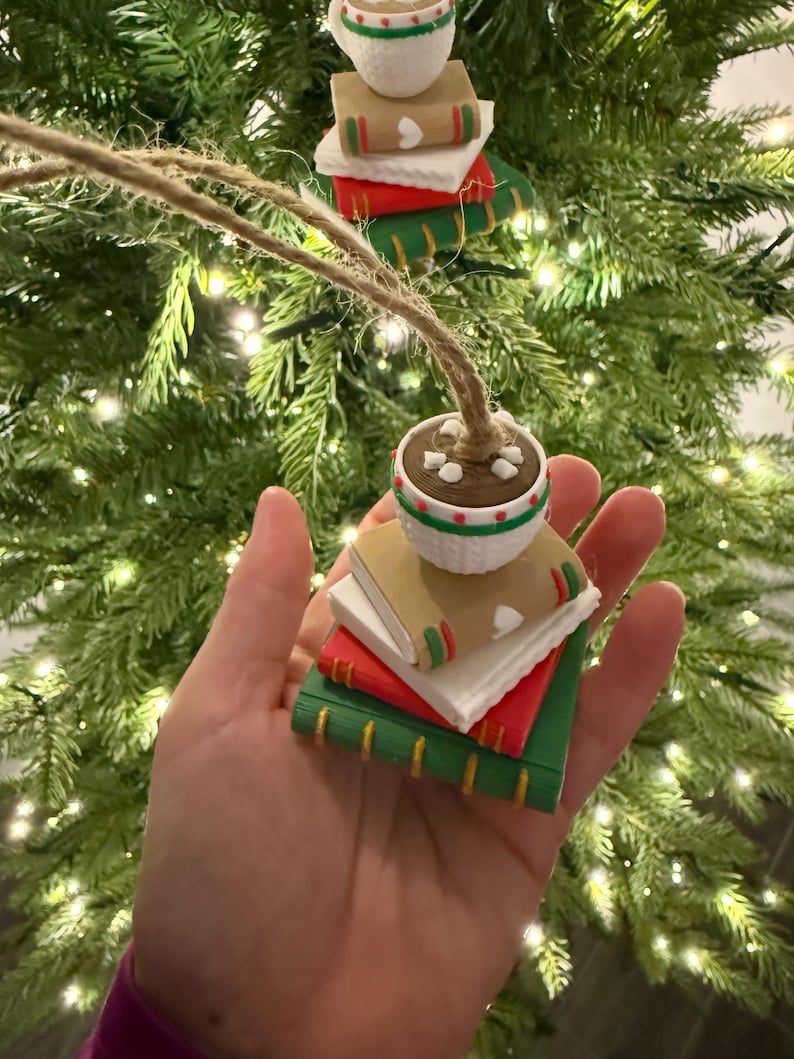 K&ouml;nnte beinhalten: Weihnachtsbaumschmuck mit einem Stapel B&uuml;cher, gekr&ouml;nt von einer Tasse Kakao. Die B&uuml;cher sind rot, gr&uuml;n und wei&szlig; mit goldfarbenen Kanten. Die Kakaotasse ist wei&szlig; mit gr&uuml;n-rotem Rand. Der Schmuck wird von einem nat&uuml;rlichen Bindfaden gehalten.
