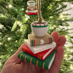 K&ouml;nnte beinhalten: Miniatur-Buchornamente mit einem kleinen Topf obenauf, an Schnur aufgeh&auml;ngt. Die B&uuml;cher sind rot, wei&szlig; und gr&uuml;n mit goldenen Akzenten. Der Topf ist wei&szlig; mit roten und gr&uuml;nen Details. Auf einem Weihnachtsbaum ausgestellt.