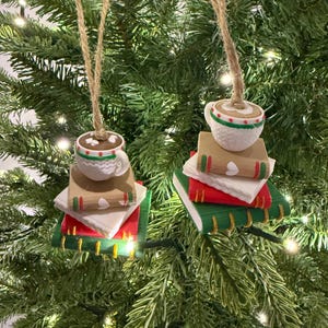 K&ouml;nnte beinhalten: Zwei Weihnachtsbaumschmuckst&uuml;cke. Jedes Ornament zeigt eine wei&szlig;e Kaffeetasse mit braunem Innenraum, auf einem Stapel von B&uuml;chern. Die B&uuml;cher sind rot, gr&uuml;n und beige, mit wei&szlig;en Seiten. Die Ornamente h&auml;ngen an Schnur.