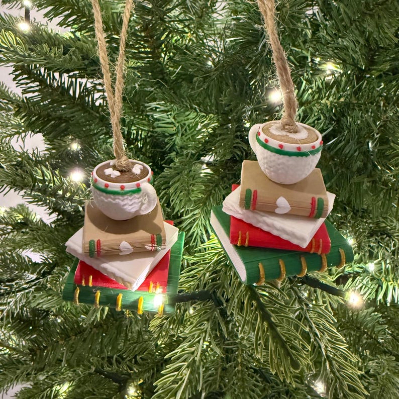 K&ouml;nnte beinhalten: Zwei Weihnachtsbaumschmuckst&uuml;cke. Jedes Ornament zeigt eine wei&szlig;e Kaffeetasse mit braunem Innenraum, auf einem Stapel von B&uuml;chern. Die B&uuml;cher sind rot, gr&uuml;n und beige, mit wei&szlig;en Seiten. Die Ornamente h&auml;ngen an Schnur.