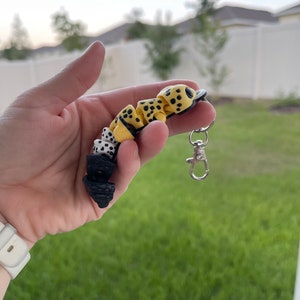 May include: A yellow, black, and white keychain with a silver clip. The keychain is made of small, round beads with a spotted pattern.