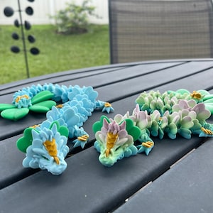 May include: Three colorful flower-shaped dragon figurines made of polymer clay. The dragons are blue, green, and pink with gold accents. The dragons are arranged on a gray table.