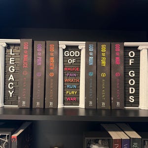 May include: A shelf display featuring a collection of books with titles like "God of Malice" and "Legacy." The books are arranged between decorative white pillars. The spines are various colors, including black, purple, and red.