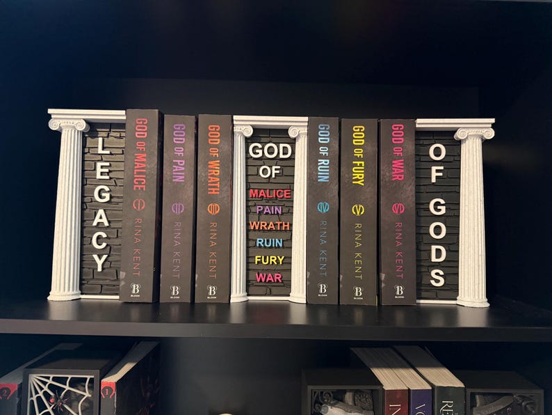 May include: A shelf display featuring a collection of books with titles like "God of Malice" and "Legacy." The books are arranged between decorative white pillars. The spines are various colors, including black, purple, and red.