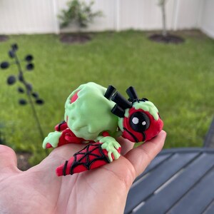 Puede incluir: Figura hecha a mano de una criatura con cabeza y patas rojas, cuerpo verde claro y detalles negros. La figura se sostiene en una mano, con un c&eacute;sped verde y una valla blanca al fondo. La criatura tiene un dise&ntilde;o de telara&ntilde;a en sus patas.