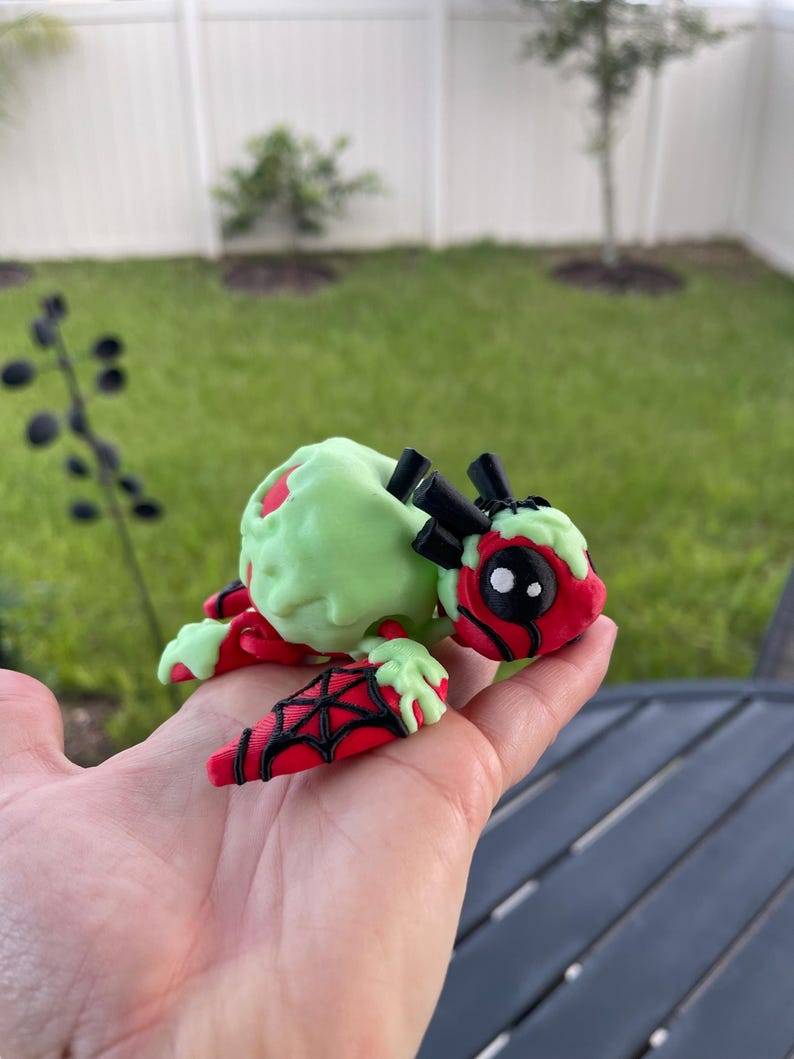 Puede incluir: Figura hecha a mano de una criatura con cabeza y patas rojas, cuerpo verde claro y detalles negros. La figura se sostiene en una mano, con un c&eacute;sped verde y una valla blanca al fondo. La criatura tiene un dise&ntilde;o de telara&ntilde;a en sus patas.