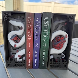 May include: A black bookend set with a gothic theme, featuring a white snake and a red hourglass on one side and a red heart on the other. The set holds three books from "The Edge of Darkness Trilogy" by Leigh Rivers.