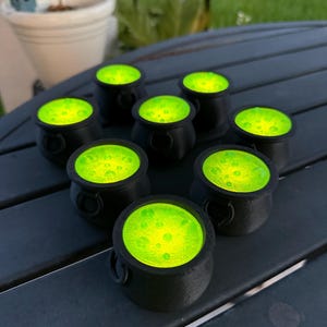 May include: Eight black cauldron-shaped candle holders with green glowing wax inside. The candle holders are arranged in a circle on a dark gray wooden surface.