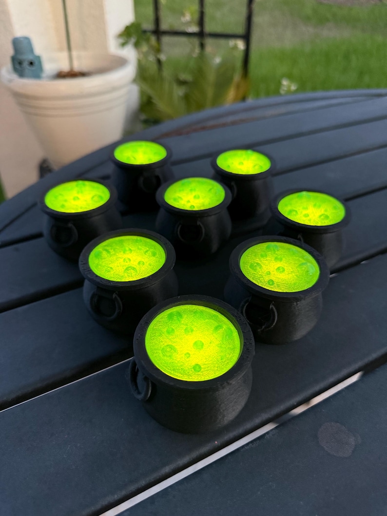May include: Eight black cauldron-shaped candle holders with green glowing wax inside. The candle holders are arranged in a circle on a dark gray wooden surface.