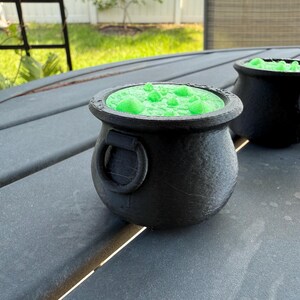 May include: Two black cauldron-shaped containers with green liquid inside. The cauldrons are sitting on a gray wooden table.
