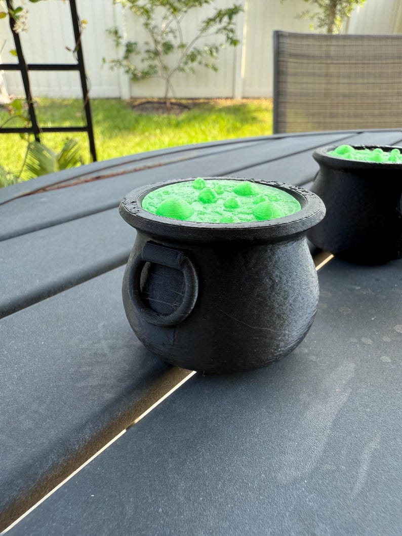 May include: Two black cauldron-shaped containers with green liquid inside. The cauldrons are sitting on a gray wooden table.